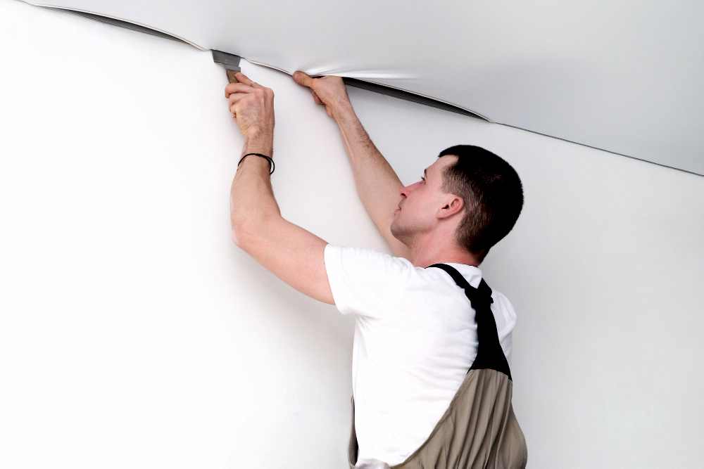 Мастер в рабочем комбинезоне и белой футболке заправляет полотно натяжного потолка в профиль