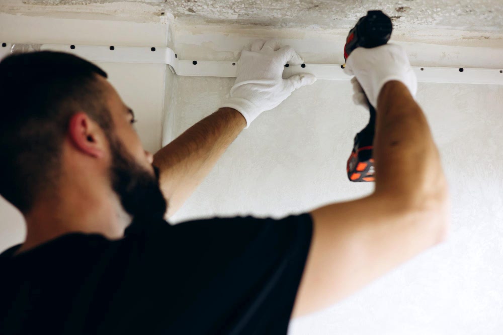 ”Мужчина в черной футболке и белых перчатках устанавливает профиль для натяжного потолка